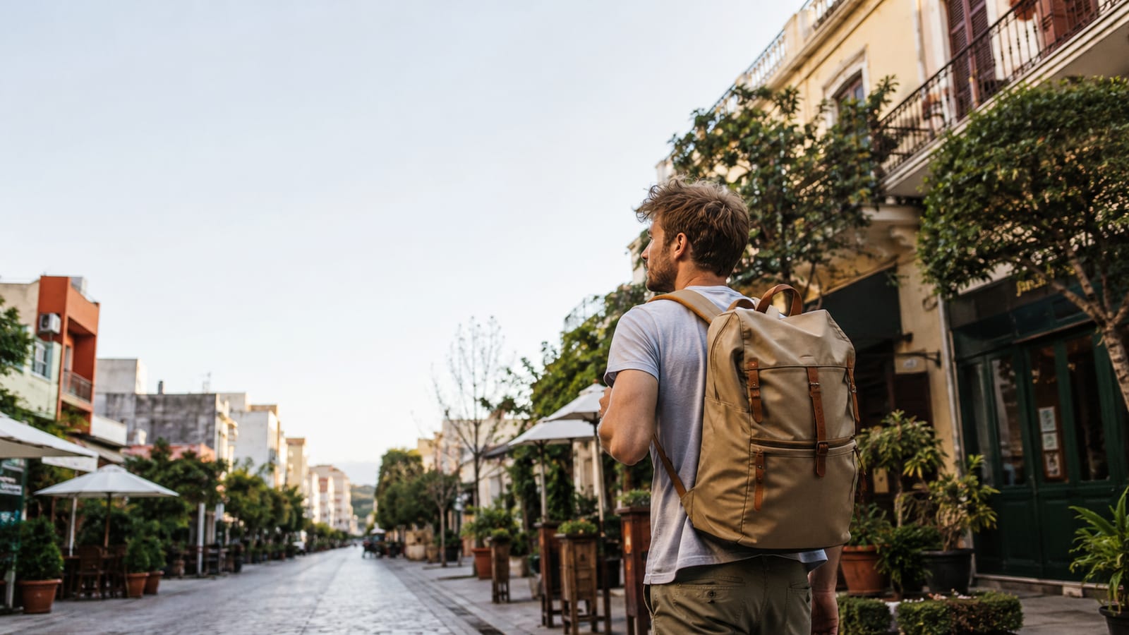 Man with tan backpack walking along a tree-lined European street at dusk