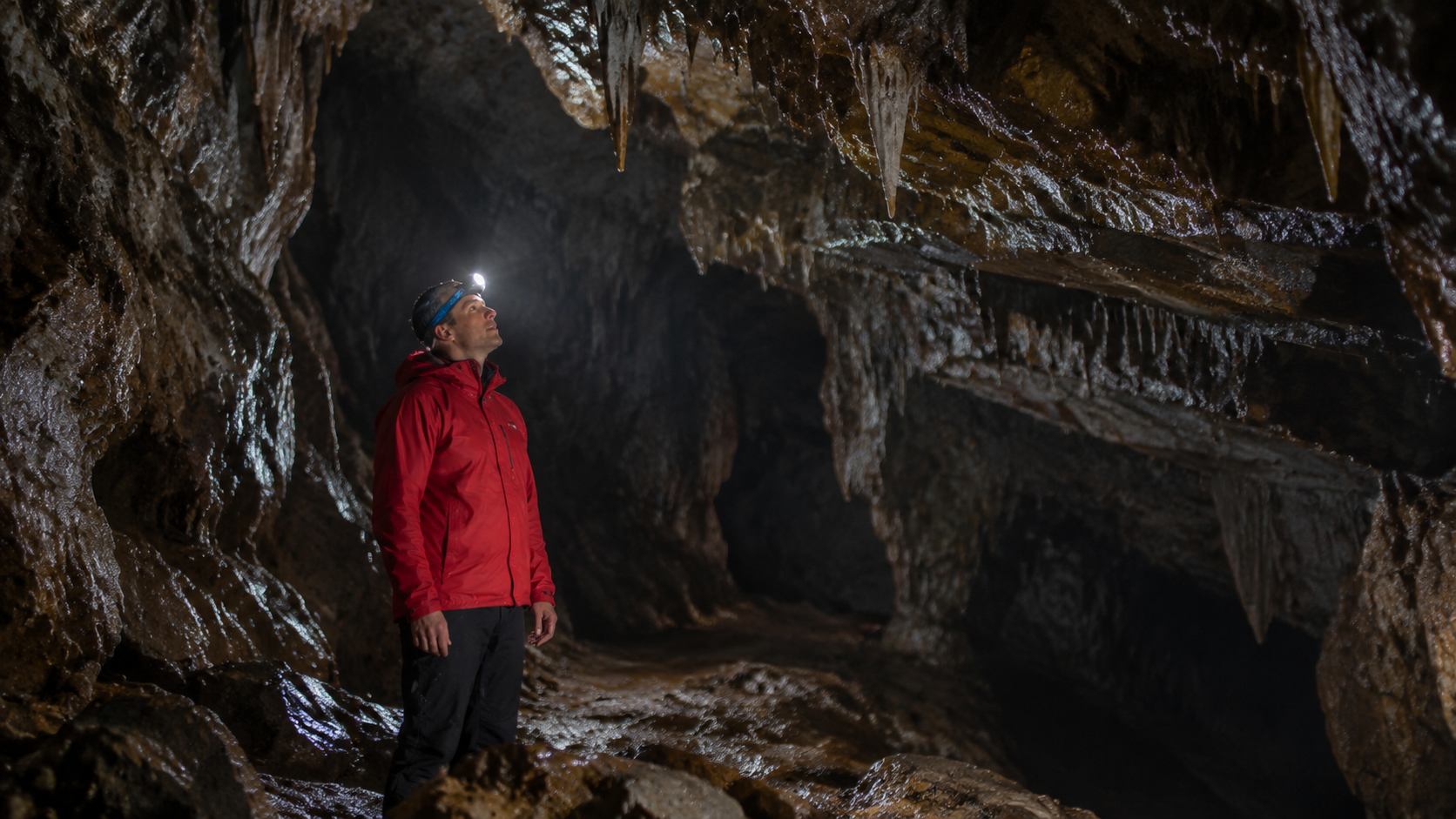 Cave explorer in headlamp and waterproof gear inside dimly lit underground cavern with damp rock surfaces