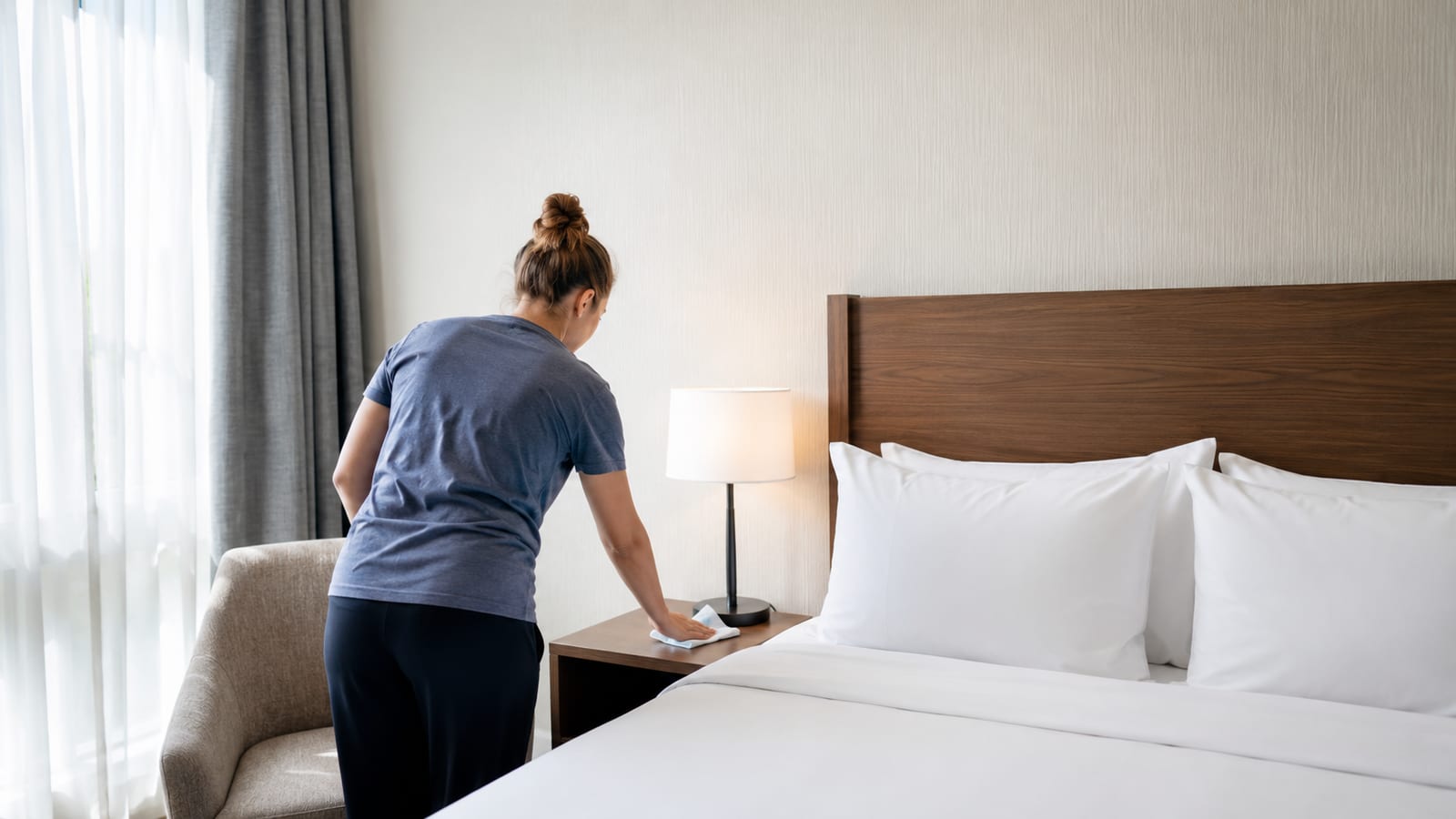 Traveler wiping hotel bedside surfaces while inspecting freshly made bedding for allergens
